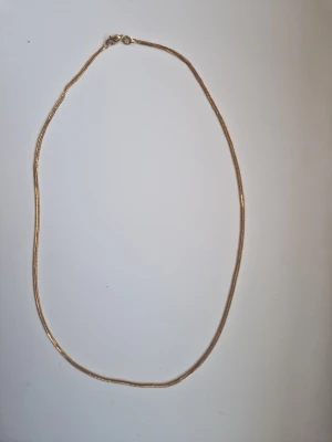 Tunt guldfärgat halsband - Säljer ett stilrent halsband i guldfärgad metall med tunn, slät kedja och klassiskt lås. Halsbandet har en enkel och minimalistisk design utan hänge eller extra detaljer. Perfekt för dig som gillar diskreta smycken i guldton.