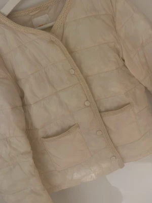 Beige pufferjacka med spetsdetaljer - Stilren beige pufferjacka från Gina tricot  med dekorativa spetsdetaljer längs kanterna och två praktiska framfickor. Jackan har tryckknappar och quiltad design som ger en mysig och trendig look. Perfekt för kalla dagar när du vill vara både varm och snygg.