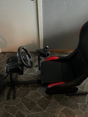 Racingsimulatorstol med ratt och pedaler - Säljer en komplett racingsimulator med svart och röd Piranha-stol, Logitech ratt, växelspak och pedaler. Stolen är sportig med skålade sidor och monterad på metallram. Perfekt för dig som vill ta bilspelandet till nästa nivå.