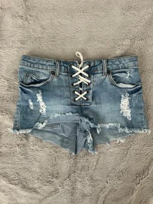 Blå jeansshorts med snörning - Säljer ett par ljusblå jeansshorts med snörning framtill och slitna detaljer. Shortsen har råa kanter och klassiska fem fickor. Perfekta för sommaren och ger en avslappnad streetstyle-look.