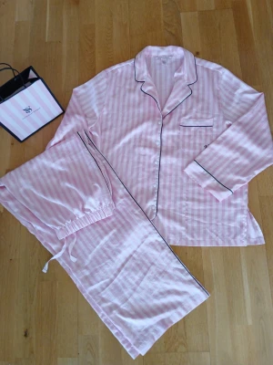 Rosa randig pyjamas från Victoria's Secret, strl L - Supermysig pyjamas i ljusrosa och vita ränder från Victoria's Secret. Setet har långärmad skjorta med svarta kanter och matchande byxor med dragsko i midjan. Sidenliknande material som känns lyxigt och skönt mot huden. Rymlig i storleken.