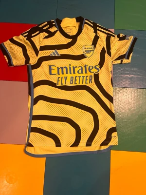 Arsenal Saka 7 bortatröja gul/svart - Cool Arsenal bortatröja från Adidas i gult med svarta mönster och blå detaljer. Tryck med Saka och nummer 7 på ryggen. Tröjan har korta ärmar och är tillverkad i lätt och ventilerande material, perfekt för fotbollsträning eller match.