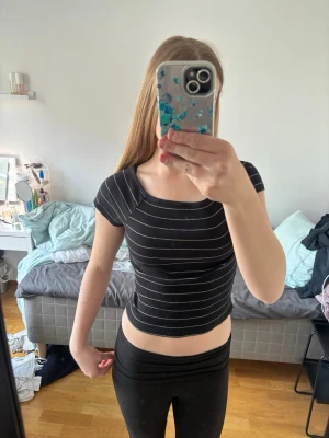 Svart croppad randig topp - Snygg svart croppad topp med vita tunna ränder och ribbad struktur. Toppen har korta ärmar och en fyrkantig ringning som ger en modern vibe. Perfekt att matcha med höga jeans eller kjol för en trendig look. Går även att ha off-shoulder.