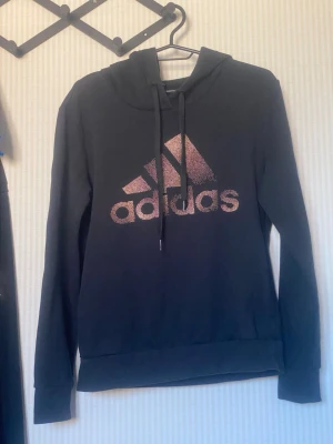 Svart hoodie från Adidas med logga - Snygg svart hoodie från Adidas med stor glittrig rosa logga på bröstet. Hoodien har huva med dragsko och långa ärmar. Tillverkad i mjukt material som känns skönt mot huden. Perfekt för en avslappnad och sportig stil.