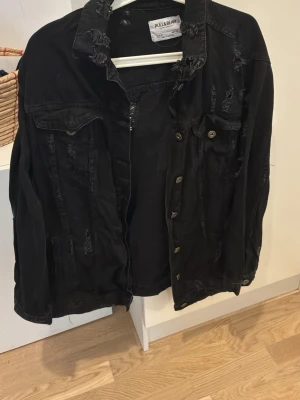 Svart jeansjacka från Pull&Bear - Cool svart jeansjacka från Pull&Bear med slitna detaljer och klassisk krage. Jackan har bröstfickor med knappar och en avslappnad passform. Perfekt för dig som gillar en edgy streetstil.