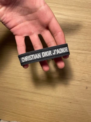 Christian Dior J'Adior armband - Snyggt vävt armband från Christian Dior i mörkgrått med vit broderad text 'CHRISTIAN DIOR J'ADIOR'. Justerbar knytning med tofsar i ändarna. Perfekt accessoar för att lyfta din outfit med en lyxig touch.