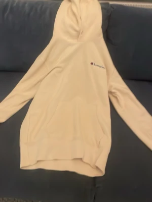 Beige hoodie från Champion - Snygg beige hoodie från Champion med broderad logga på bröstet. Klassisk modell med huva och känguruficka framtill. Mjuk insida och ribbade muddar vid ärmslut och nederkant. Perfekt för en avslappnad och trendig stil.