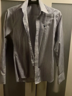 Randig skjorta från Ralph Lauren - Klassisk skjorta med smala blå och vita ränder från Ralph Lauren. Skjortan har lång ärm, krage och broderad logga på bröstet. Perfekt för en stilren look och enkel att matcha med jeans eller chinos.