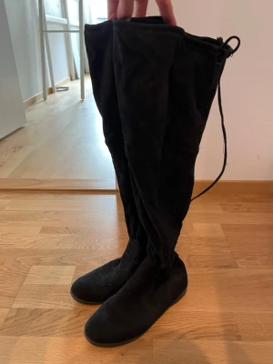 Svarta overknee stövlar i mocka-look - Snygga svarta overknee stövlar med snörning upptill. Gjorda i ett mjukt material med mocka-känsla och platt sula. Perfekta för att lyfta vilken outfit som helst och ger en cool vibe. Benen är mjuka, alltså inte styva och håller inte upp sig själva. Välanvända. 