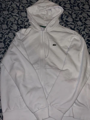 Vit hoodie från Lacoste med dragkedja - Säljer en clean vit hoodie från Lacoste med dragkedja och klassisk krokodillogga på bröstet. Hoodien har huva med dragsko och fickor framtill. Perfekt för dig som gillar stilren streetwear och vill ha något som funkar året runt.