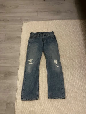 Levi's raka blå jeans med slitningar - Säljer ett par klassiska blå jeans från Levi's med raka ben och slitna detaljer på båda benen. Jeansen har fem fickor och en traditionell knappgylf. Perfekta för en avslappnad och trendig look.