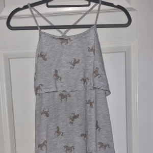 Grå långklänning med hästmönster - En grå långklänning med smala axelband och ett hästmönster. Klänningen har en lös passform och är ärmlös med en volangdetalj över bysten. Från H&M i stl 146/152