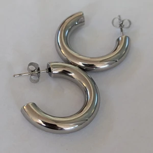 Silverfärgade örhängen - Ett par silverfärgade örhängen i en rund, tjock design. Örhängena har en slät och blank yta och är av typen stiftörhängen med fjärilslås.aldrig använt, granska lite tunga, rostfritt stål 