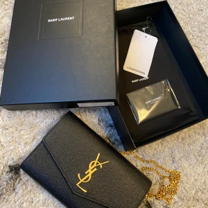 Saint Laurent Uptown clutch - - Svart - Höjd 12 cm - Bredd 19 cm - Djup 2,5 cm - Avtagbar guld kedja - Kommer med en korthållare - Made in Italy - 100% kalvskinn   Köpt på Luisaviaroma. Kvitto finns.   Nypris ca 13000 kr