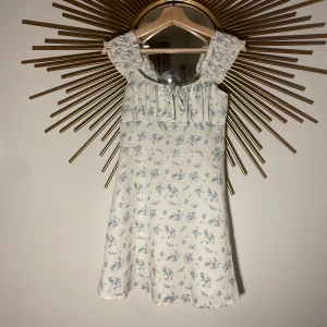 Blommig sommarklänning med volangdetaljer - En jättesöt sommarklänning med blommigt mönster i blått och grönt på en vit bakgrund. Klänningen har volangdetaljer vid axelbanden och en knytning framtill. Sitter snyggt på och är helt oanvänd!🌸🙌