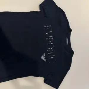 En svart t-shirt från Emporio Armani med korta ärmar. T-shirten har texten 'EMPORIO' och Emporio Armanis logotyp tryckt på framsidan i en glittrig stil.