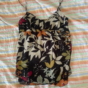 Blommig linnetopp från H&M - En blommig linnetopp från H&M med tunna axelband. Toppen har ett mönster i svart, beige, orange, röd och grön. Den är ärmlös och har en lös passform.