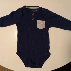 Marinblå body strl. 56 - Marinblå body med dekorativa knappar i storlek 56 från Petite mini. I fint begagnat skick, inga hål eller fläckar. Något blekare än ny och lite färg har skavts bort från knapparna vid axeln.  Tvättad i parfymfritt tvättmedel och utan sköljmedel.