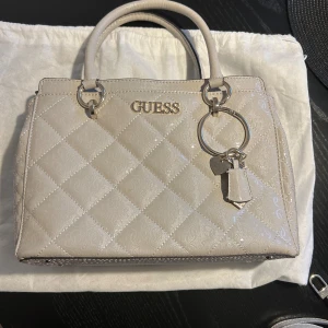Guess väska i nyskick - Perfekt väska för både vardagen & speciella tillfälle. Bild 1 visar den rätta färgen, beige. Använda vid 2 tillfälle, som ny. Flera praktiska fack, säker dragkedja & justerbart axelband ingår. Ca. 23x33 cm.  Säljer även en matchande plånbok.