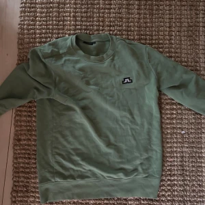 Grön tröja från J.Lindeberg - En grön tröja från J.Lindeberg med rund halsringning och långa ärmar. Tröjan har en enkel design med en liten logotyp på bröstet.