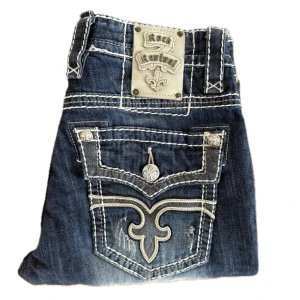 Riktigt feta drain rock revival bootcut baggy jeans - Fett drain o snygga rock revival jeans i super bra skick, knappt använda. Har lite distressing på låren o bakfickan, vilket är en del av designen. Midja: 41 cm. Längd: 113 cm. Benöppning: 22 cm. Ställ gärna frågor, priset kan diskuteras.