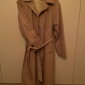 Trenchcoat begie xs Nyskick! - Supersnygg enkel trenchcoat från Indiska. Inköpt våren 2023. Använd fåtal gånger. Rök och djurfritt. Snygga knappar. Slag i ryggen. Perfekt begie färg (ej gul, ej grön, ej blek).