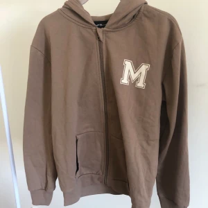 Zip up tröja - Brun zip up tröja med ett tryck av ett M på sidan, använd någon gång, bra skick
