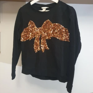 Jultröja med glittrig rosett  - Svart jultröja med rosett i glitter med bronsfärgade paljetter. Sparsamt använd. Perfekt nu till vintern! 💛 OBS katt finns i hemmet! Passar XS-M beroende på vilken passform man vill ha 