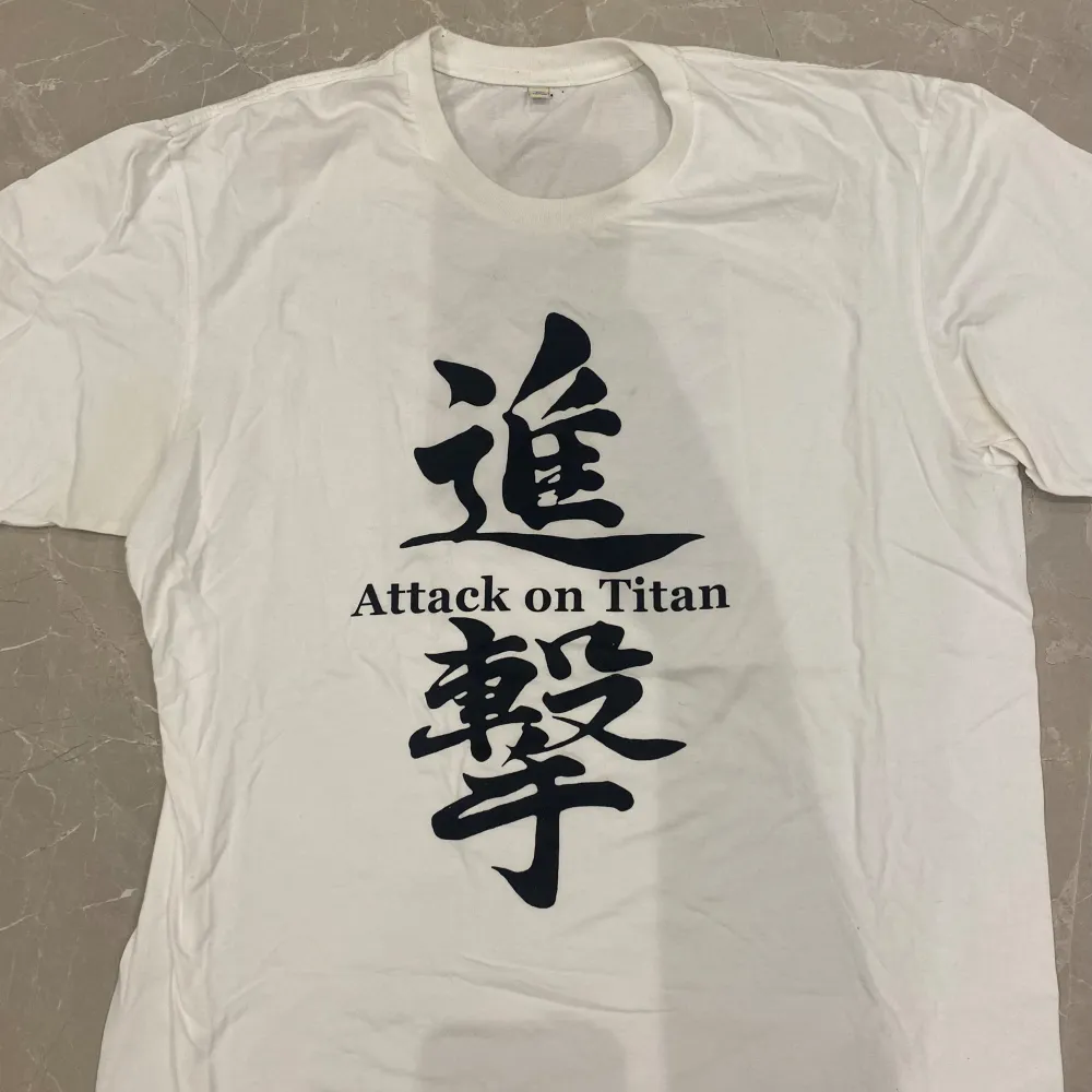  En vit t-shirt och med den animerade ”Attack on titan”  loggan på fram och baksidan . T-shirts.