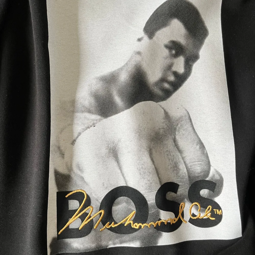 En svart tröja från Hugo Boss med ett stort tryck av Muhammad Ali på framsidan. Tröjan har långa ärmar och en rund halsringning. Trycket är i svartvitt med texten 'BOSS' och Muhammad Alis signatur i guld.. Tröjor & Koftor.