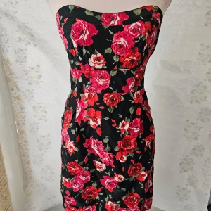 Blommig klänning från Cache Cache - En elegant axelbandslös klänning från Cache Cache med ett vackert blommönster i rött och rosa på en svart bakgrund. Klänningen har en tight passform och är perfekt för festliga tillfällen.