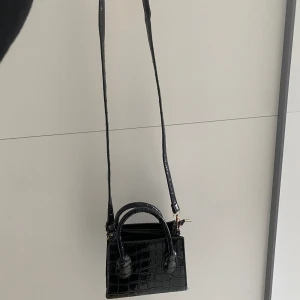 Väska - En lite mindre handväska som kan bäras med det långa snöret eller att man bara håller i den.  En lite mindre skada på sidan där snöret är fäst då den lossnat, men har ”fixats” med att sy ihop det.