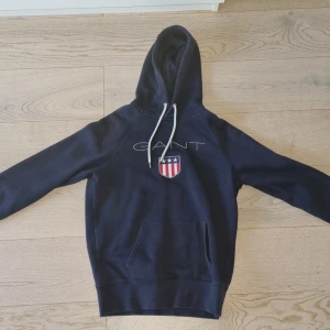 Mörkblå hoodie från GANT - Säljer en snygg och bekväm mörkblå hoodie från GANT. Den har en stor ficka på magen och en luva med vita snören. Perfekt för höst och vinter!