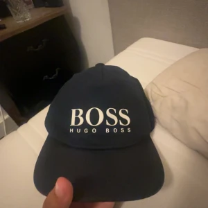 Svart och blå keps från Hugo Boss - Säljer två snygga kepsar från Hugo Boss i färgerna svart och blå. Båda kepsarna har en klassisk design med 'BOSS' och 'HUGO BOSS' tryckt i vitt på framsidan. Perfekt för både vardag och sportiga aktiviteter. Kepsarna är i bra skick och redo för en ny ägare!