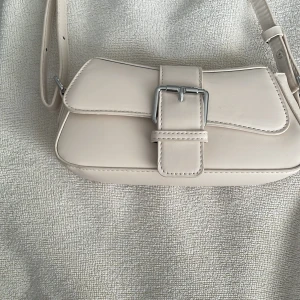 Beige axelväska med spänne - Säljer en snygg beige axelväska. Väskan har ett stort spänne på framsidan och en justerbar axelrem. Perfekt för både vardag och fest! 👜✨