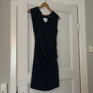 Mörkblå ärmlös klänning - Säljer en elegant mörkblå klänning som är ärmlös och har en v-ringad hals. Klänningen har en draperad detalj på sidan som ger en snygg och stilren look. Perfekt för fest eller andra speciella tillfällen. Klänningen är i nyskick och passar bra till både sommar och vår.