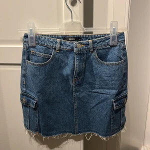 Blå jeanskjol från Bik Bok - En blå jeanskjol från Bik Bok i storlek 38. Kjolen har en rå kant nedtill och är utrustad med fickor både fram och bak samt på sidorna. Den har en knapp och dragkedja framtill.