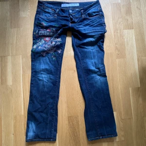 Mörkblå jeans från Ed Hardy - Säljer ett par mörkblå jeans från Ed Hardy med coola rhinestone detaljer på vänster ben. Jeansen har en straight/bootcut fit och är i bra skick. Perfekta för en y2k look. Passar både till vardags och fest!