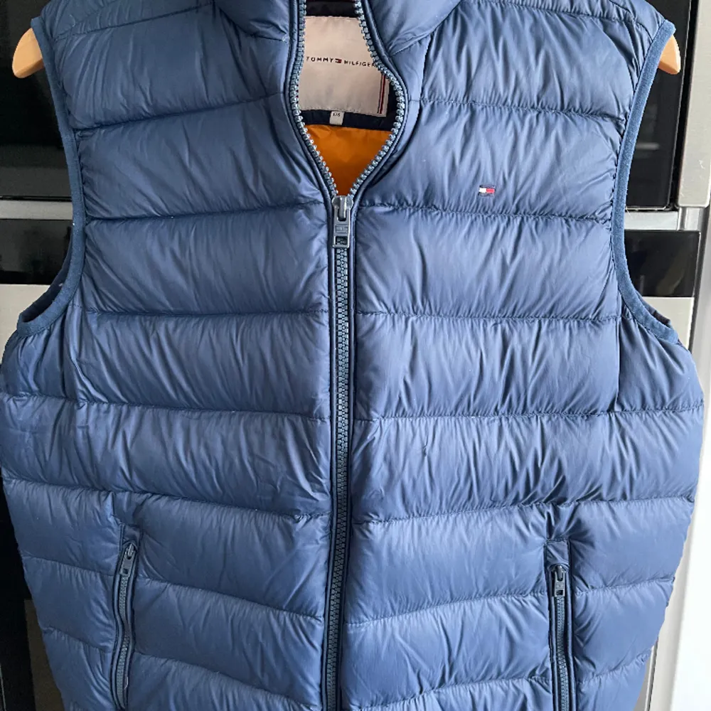 Marinblå dunväst från Tommy Hilfiger med orange foder. Knappt använd. Inget slitage. Västen går att lägga in i en ficka och bära med sig på ett smidigt sätt runt midjan. 15-17 år. 176 cm. Jackor.