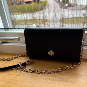 Svart axelremsväska väska från Tory Burch. Burch - Är i jättefint skick och är köpt på Crisma i Örebro år 2015. Väskan är 19,5 cm bred och 13 cm hög. Man får plats med till exempel mobiltelefon och betalkort i väskan. 