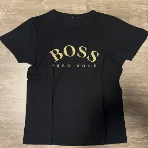 Säljer en svart Hugo Boss t-shirt i storlek M med guldfärgad logo på bröstet. T-shirten är i mycket bra skick och tillverkad av mjukt och bekvämt material. 