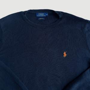 VARAN KAN KÖPAS DIREKT PÅ VÅR HEMSIDA, ettmarke.se MÄRKE: Ralph Lauren. FÄRG: Mörkblå/svart med orange logga. STORLEK: M. AXELBREDD (SÖM TILL SÖM RAKT ÖVER): 45 cm. TOTALLÄNGD (FRÅN KRAGEN): 70 cm. ÄRMLÄNGD (ÄRMHÅLAN TILL MUDDSLUT): 56 cm. 