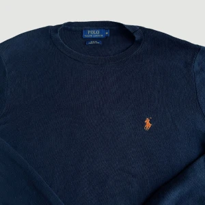 Ralph Lauren - Stickad Tröja - Storlek M - VARAN KAN KÖPAS DIREKT PÅ VÅR HEMSIDA, ettmarke.se MÄRKE: Ralph Lauren. FÄRG: Mörkblå/svart med orange logga. STORLEK: M. AXELBREDD (SÖM TILL SÖM RAKT ÖVER): 45 cm. TOTALLÄNGD (FRÅN KRAGEN): 70 cm. ÄRMLÄNGD (ÄRMHÅLAN TILL MUDDSLUT): 56 cm. 