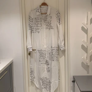 Vit långklänning med textmönster - En vit långklänning med svart textmönster. Klänningen har långa ärmar och en krage. Den är knäppt framtill med knappar och har en lös passform.Helt nytt aldrig använd