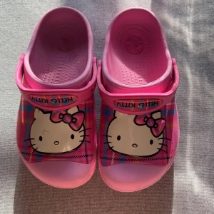 Hello kitty skor - Ett par rosa Crocs med Hello Kitty-motiv. Skorna har en justerbar rem och ventilationshål på sidorna. Hello Kitty-motivet är placerat på ovansidan av skorna och remmen har texten 'Hello Kitty'. de är i strl 3 som motsvarar ungefär 35 kanske 36, super gulliga, lite använda som man kan se på bild 4 annars fina original pris 600kr 