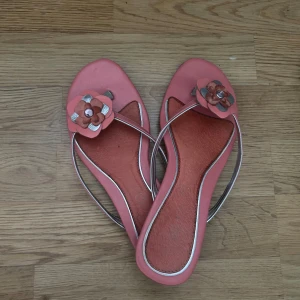 Rosa sandaler med blommig detalj - Ett par rosa sandaler med en dekorativ blomma på remmen. Sandalerna har en öppen tå och en tunn rem som går över foten.