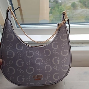 Grå axelväska från Guess - En elegant grå axelväska från Guess med ett mönster av G-logotyper. Väskan har en guldfärgad kedja och dragkedja samt en justerbar axelrem.