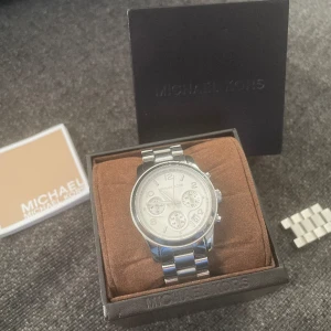 Michael kors klocka  - En silvrig Michael kors klocka. Någon ytlig repa finns då den är använd ju men inget som störs. Extra länkar o Ask medföljer! Batteri byte behövs då den legat i lådan länge.  Självklart äkta!!