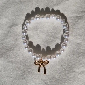 Pärlarmband med Guldrosett - °●☆•○☆● Gjort av elastisk tråd, vita pärlor, och rosetthänge. Minimalistisk och söt accessoar som passar till alla tillfällen. Perfekt som gåva ♡ Är i fint skick, helt ny, och har inga skador.
