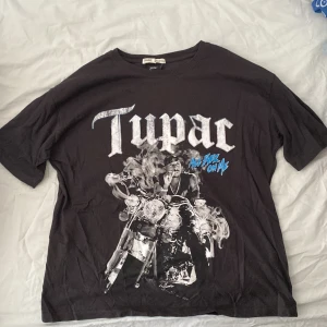 Svart T-shirt med Tupac-tryck från Pull&Bear - En svart T-shirt från Pull&Bear med ett stort tryck av Tupac på framsidan. Texten 'Tupac' och 'All Eyez On Me' är tryckt i vitt och blått. T-shirten har korta ärmar och en rund halsringning.